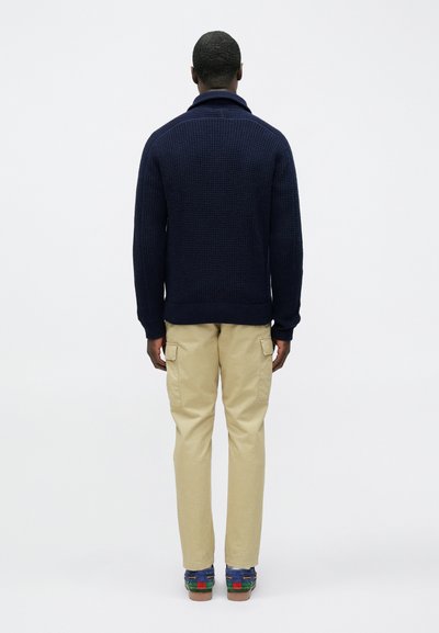 Tmavomodrý pletený sveter s vysokým golierom, nosený s khaki cargo nohavicami a farebnými slip-on topánkami. Tkanina pôsobí textúrovaným a hrubým dojmom.
