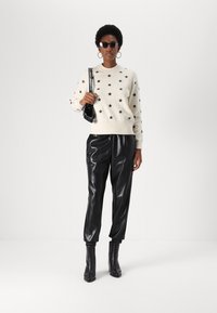 Pull en ivoire avec un motif d'étoiles noires, pantalon en simili cuir noir brillant et bottines noires. Les accessoires comprennent des lunettes de soleil et un petit sac.
