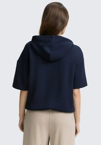 Tengerészkék kapucnis pulóver rövid ujjakkal, texturált anyagból és laza szabással, bézs nadrággal párosítva, amelynek hátsó zsebei vannak.