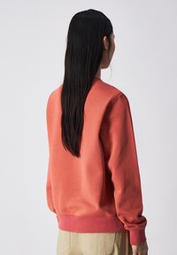 Sudadera color coral con cuello redondo y puños acanalados. Textura de tela suave, corte relajado y una cinturilla que coincide en color con los puños.