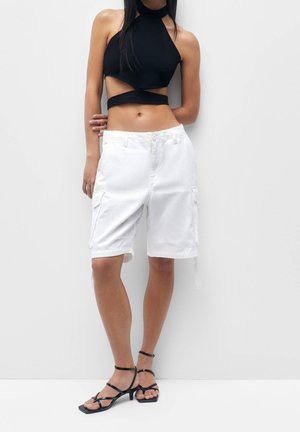 Shorts cargo blancs avec poches latérales, accompagnés d'un top halter noir. La tenue présente un design court et des sandales noires à lanières.