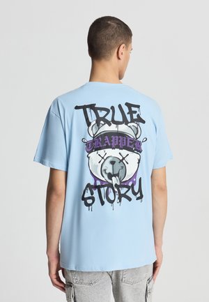 T-shirt in cotone azzurro chiaro con grafica di un orso sul retro e testo nero e viola che recita "TRUE TRAPPER STORY." Maniche corte, vestibilità comoda.
