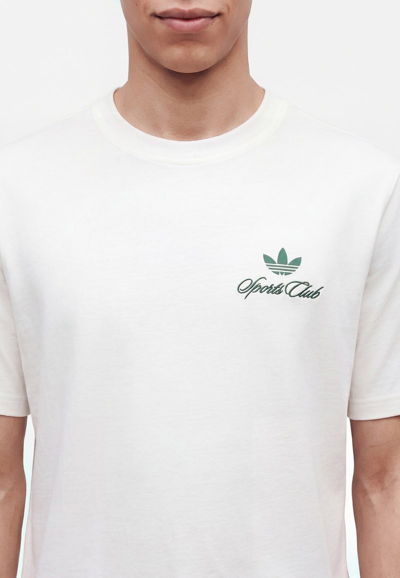Person trägt ein weißes T-Shirt mit grünem "Sports Club"-Logo und dreiblättrigem Emblem auf der linken Brustseite.