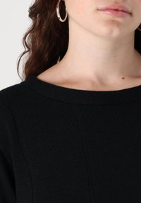 Maglione nero lavorato a maglia con un ampio scollo rotondo e dettagli in sottile texture, incorniciato da capelli lunghi e mossi e orecchini a cerchio in argento.