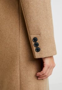 Cappotto in misto cammello con taglio dritto, caratterizzato da tre bottoni neri sulla manica e una finitura liscia e strutturata.