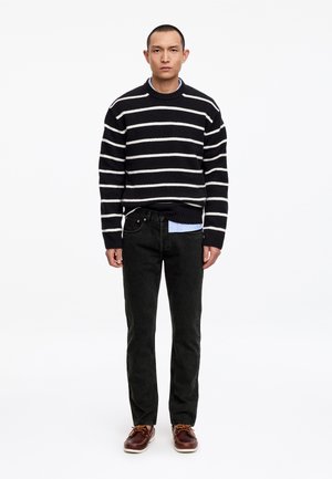 Hombre de pie mirando hacia adelante, vestido con un suéter a rayas blancas y negras, jeans negros y zapatos marrones con cordones, sobre un fondo blanco.