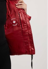 Chaqueta acolchada roja con una textura suave, presenta un logo de Khujo, un bolsillo interior y un dobladillo con cordón para mayor ajustabilidad.
