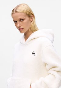 Valkoinen fleece-huppari, jossa on suuri huppu, kengurupocket ja pieni musta brodeerattu logo rinnassa. Pehmeä, teksturoitu kangas.