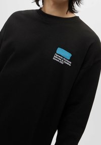 Osoba ubrana w czarną bluzę z kapturem z niebieskim prostokątem i białym napisem na klatce piersiowej: "Pigments Worldwide Biennale du Système Sonore 2022".