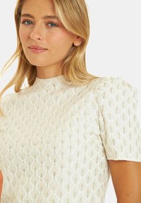Biała bluzka o teksturowanej powierzchni z wzorem w diamenty, krótkimi bufkami oraz wysokim dekoltem z subtelnymi metalicznymi akcentami.