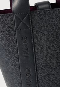 Geantă tote din piele neagră texturată, cu mânere netede negre contrastante. Prezintă un logo „HUGO” embossat pe bretea. Interiorul are o captuseală burgundy.