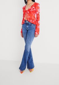 Mujer vistiendo una blusa larga de manga larga con flores rojas y volantes, jeans de campana de tiro alto en azul, y tacones abiertos y coloridos, de pie sobre un suelo blanco.