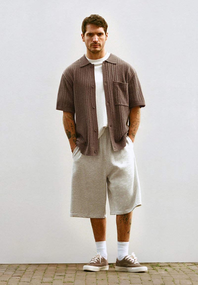Homme portant un cardigan marron à manches courtes par-dessus une chemise blanche, un short gris clair au niveau des genoux, des chaussettes blanches et des baskets marron, debout sur un sol en briques.