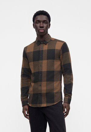 Jeune homme portant une grande chemise à carreaux marron et noire boutonnée et un pantalon noir, debout devant un fond clair uni.