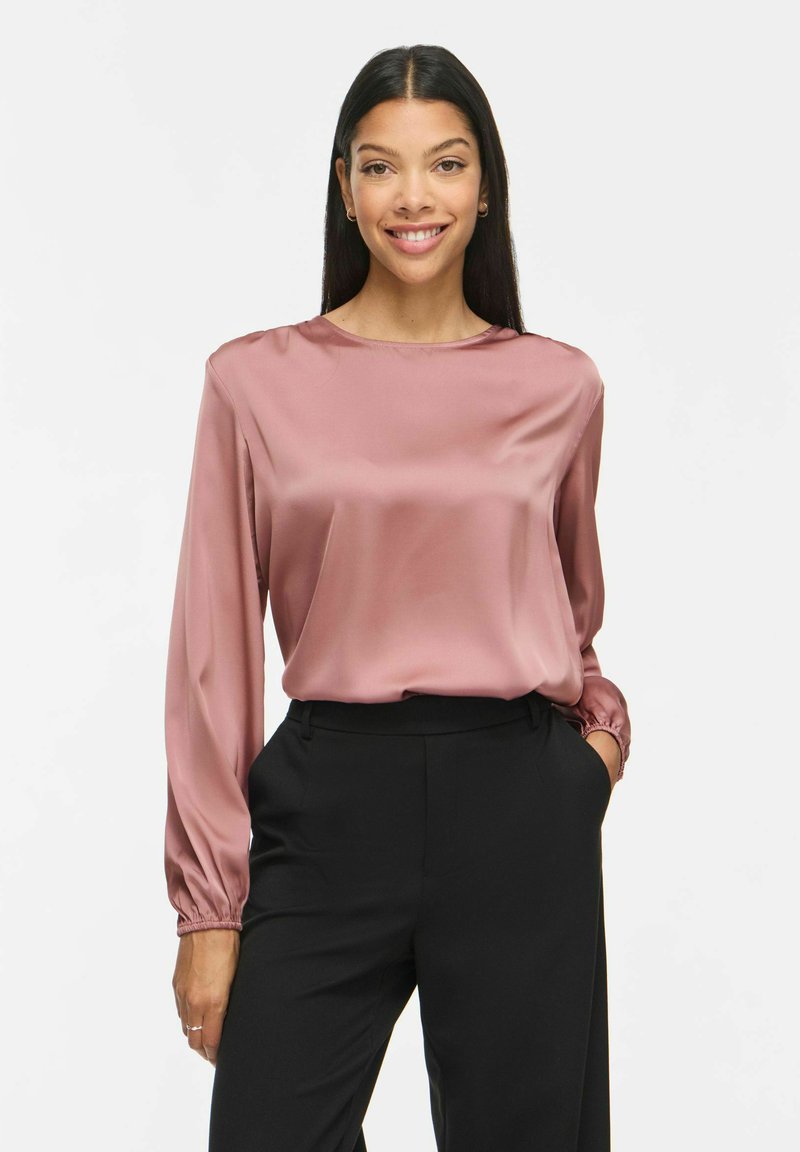 Jeune femme souriante portant un chemisier rose en satin à manches longues et un pantalon noir, une main dans la poche sur fond blanc.