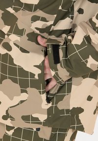 Beige og grøn jakke med camouflage-mønster og rutemønster. Har en lynlåslomme og en velcro-lukning på ærmet.