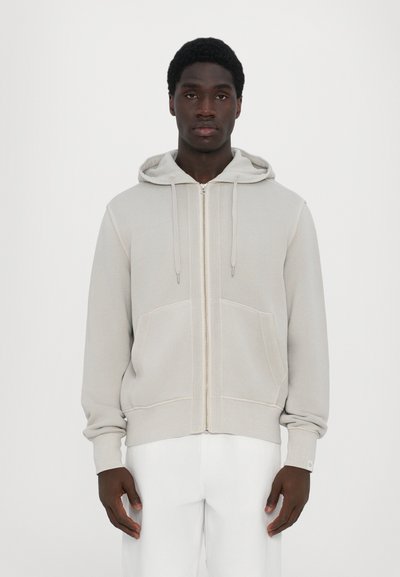 Sweat à capuche zippé gris clair avec une poche frontale, un cordon de serrage à la capuche et des poignets côtelés. Le tissu semble doux avec une texture lisse.