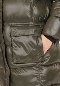 Olivegrön puffjacka med en glänsande, quiltad yta, stora framfickor och en kroppsnära passform runt torso och ärmar.