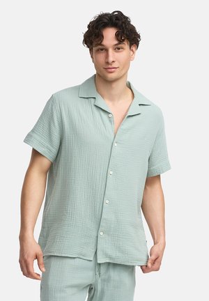 Camisa verde clara de manga corta hecha de tela texturizada, con un cierre de botones en la parte delantera, cuello con muescas y un discreto logo de la marca en el pecho.