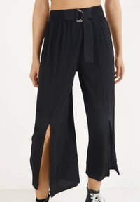 Black pleated wide-leg pants with a belted waistband, featuring side slits and a textured fabric for added detail.