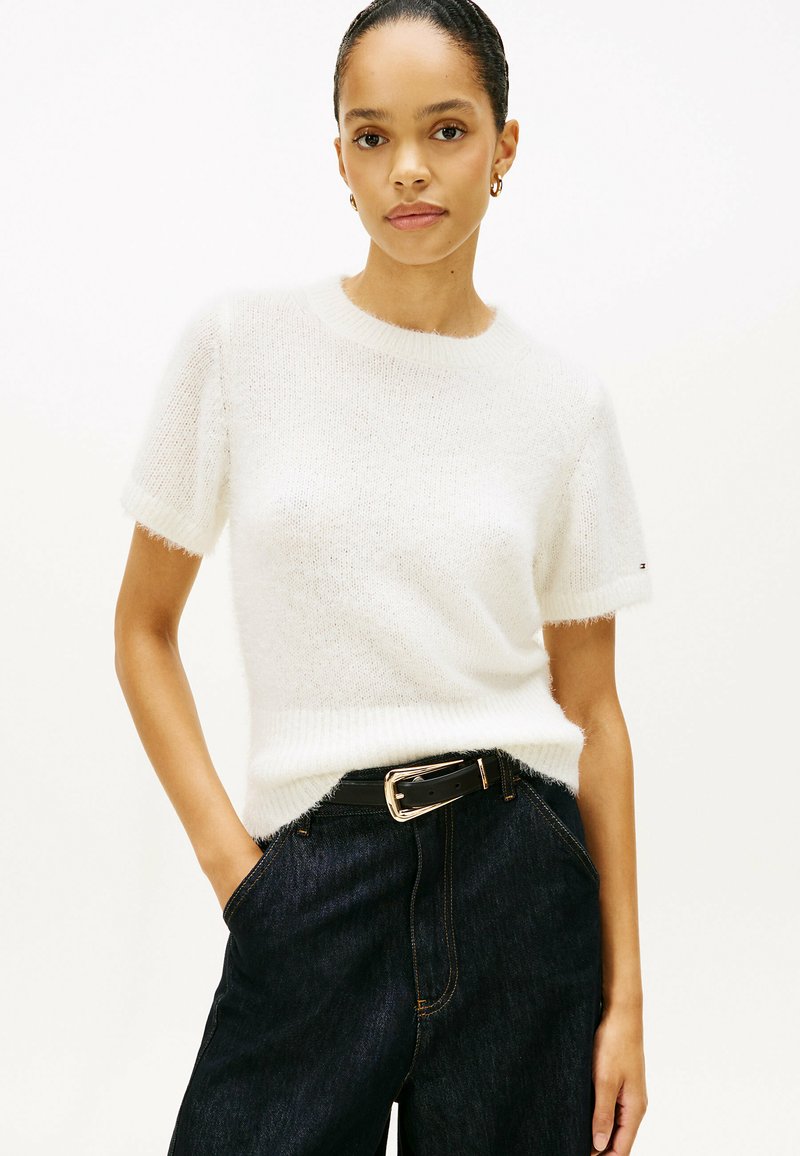 Vrouw met handen in de zakken van donkere jeans, gekleed in een wit, kortärmelig, gebreid truitje en gouden oorringen, staand tegen een witte achtergrond.