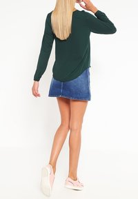 Blusa verde a maniche lunghe abbinata a una gonna di jeans blu. La blusa è liscia, mentre la gonna presenta una classica texture in denim. Sneakers chiare completano l'outfit.