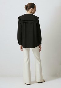 Maglietta nera a maniche lunghe con un grande colletto strutturato e dettagli testurizzati, abbinata a pantaloni bianchi svasati e scarpe beige.