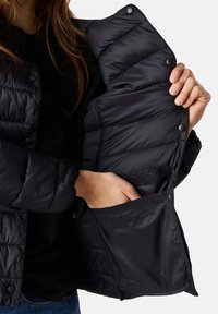 Giacca isolante nera con un design trapuntato, dotata di bottoni a pressione e una tasca interna. Il materiale appare liscio e leggermente lucido.