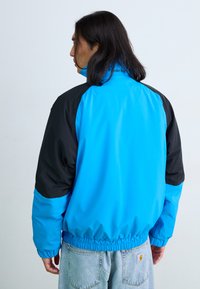 Blau- und schwarze Windjacke mit elastischem Saum, hochschließendem Kragen und Kontrastbesätzen, gefertigt aus einem glatten, leichten Material.