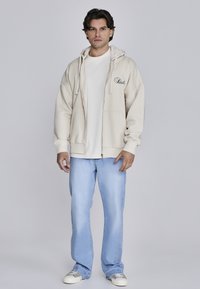 Sudadera cremosa con cremallera y cordones, que cuenta con un bolsillo frontal y un logo. Combinada con una camiseta blanca y jeans rectos de color azul claro.