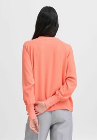Maglia a maniche lunghe color corallo realizzata in tessuto morbido, con vestibilità rilassata, maniche raglan e polsini a coste. Abbinata a pantaloni grigi chiari.