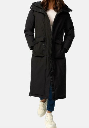 Femme portant un long manteau d'hiver noir à capuche avec des poches avant, jean bleu, baskets blanches, debout les mains dans les poches.