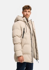 Chaqueta acolchada beige con capucha, tejido texturizado, bolsillos con cremallera y detalles en negro. El cierre frontal cuenta con broches y una cremallera.