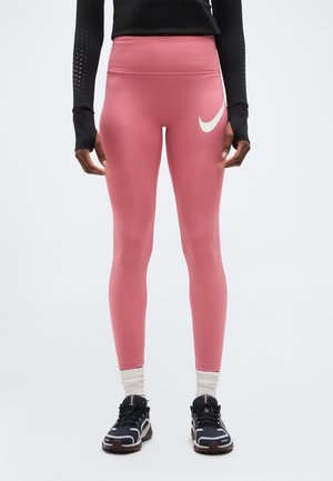 Persoon die een roze sportlegging met hoge taille en een wit Nike-logo draagt, een zwart shirt met lange mouwen, witte sokken en zwarte hardloopschoenen, staand.