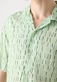 Chemise vert clair à manches courtes boutonnée avec un motif abstrait de tirets verticaux noirs et blancs, portée par une personne avec un cou et une épaule visibles.