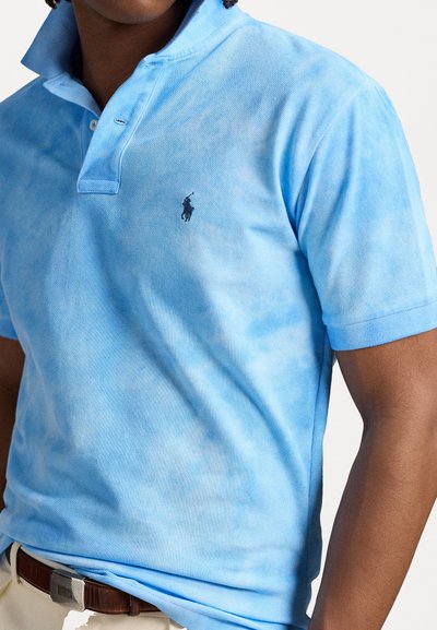 Modrá tie-dye polo košile vyrobená z bavlny, s límcem na dva knoflíky, krátkými rukávy a malým vyšitým logem na levé hrudi.