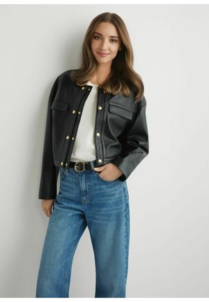 Młoda kobieta z długimi brązowymi włosami, ubrana w czarną skórzaną kurtkę, biały top, niebieskie dżinsy i czarny pasek, stojąca przy białej ścianie.