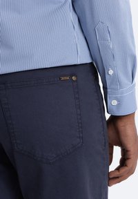 Pantalon bleu marine en tissu doux, avec une poche arrière portant une étiquette en métal et une texture subtile. La chemise est bleu clair avec de fines rayures.