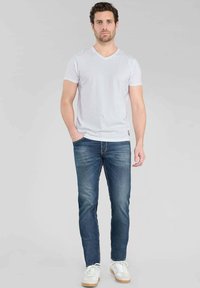 T-shirt bianco a V abbinato a jeans di media tonalità. I jeans sono a vestibilità slim con un classico design a cinque tasche, indossati con sneakers bianche.