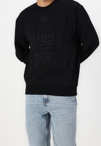 Black sweatshirt featuring embossed logo and text, long sleeves, round neckline, and ribbed cuffs, paired with light blue jeans.