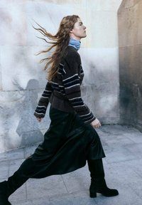 Maglione a righe marroni e blu, gonna di pelle nera e stivali neri al ginocchio. Capelli lunghi che fluiscono, mentre cammina contro uno sfondo di pietra chiara.