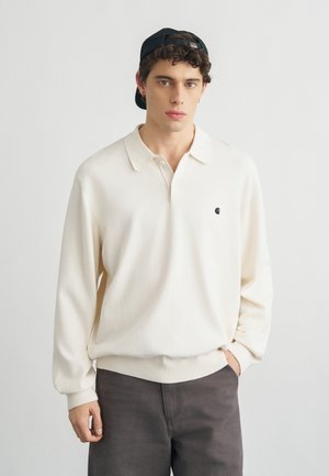 Giovane uomo con capelli ricci che indossa un berretto nero a rovescio, una polo crema a maniche lunghe e pantaloni scuri, in piedi davanti a uno sfondo chiaro e semplice.