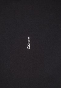 Tissu noir à texture lisse avec un logo vertical blanc "HUGO" en police moderne centré sur la surface.