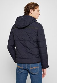 Marineblaue Steppjacke mit abnehmbarem Kapuzen, horizontaler Steppstruktur und kontrastierendem schwarzen Streifen über den Schultern. Getragen mit blauen Jeans.