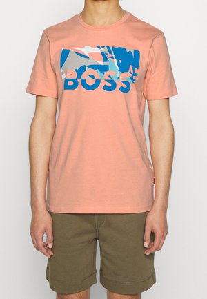 Personne portant un t-shirt à manches courtes pêche avec un motif abstrait bleu et gris et le texte « BOSS », associé à un short vert olive.