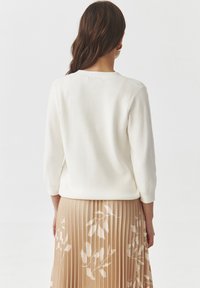 White ribbed sweater with three-quarter sleeves, paired with a beige pleated skirt featuring a floral pattern. Smooth texture, minimal design.