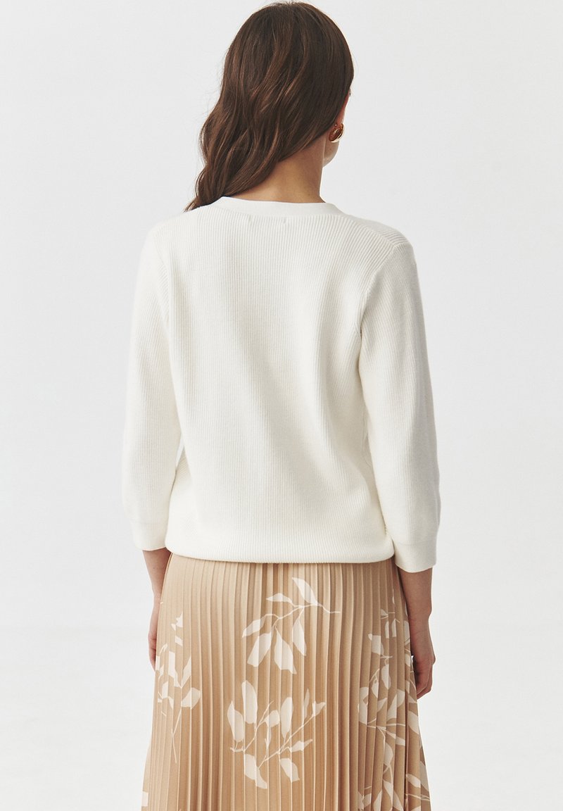 Biała sweter z ribbingu z rękawami o długości trzech czwartej, w połączeniu z beżową spódnicą plisowaną zdobioną kwiatowym wzorem. Gładka tekstura, minimalny design.