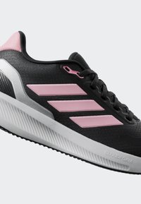 Chaussure de sport noire avec des accents roses, tige en maille texturée, intermédiaire rembourré blanc et semelle extérieure en caoutchouc. Design à trois bandes remarquable.