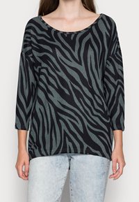 Schwarze und grüne Bluse mit Zebra-Streifen, weitem Ausschnitt und Dreiviertelärmeln, aus einem weichen, strukturierten Stoff gefertigt.