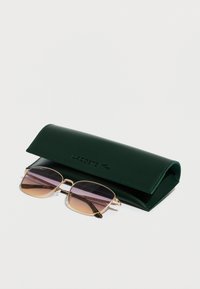 Hus�ă verde din piele cu logo-ul LACOSTE, care conține ochelari de soare cu ramă aurie și lentile degrade mov, având o formă pătrată și accente delicate.